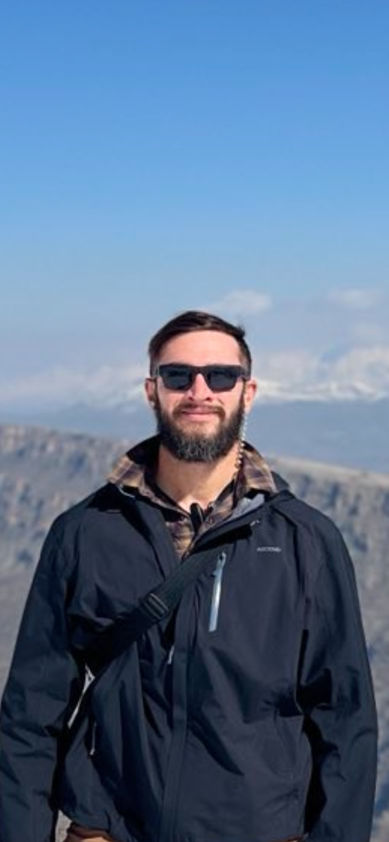 Man wearing sunglasses and a dark jacket with mountains in the background