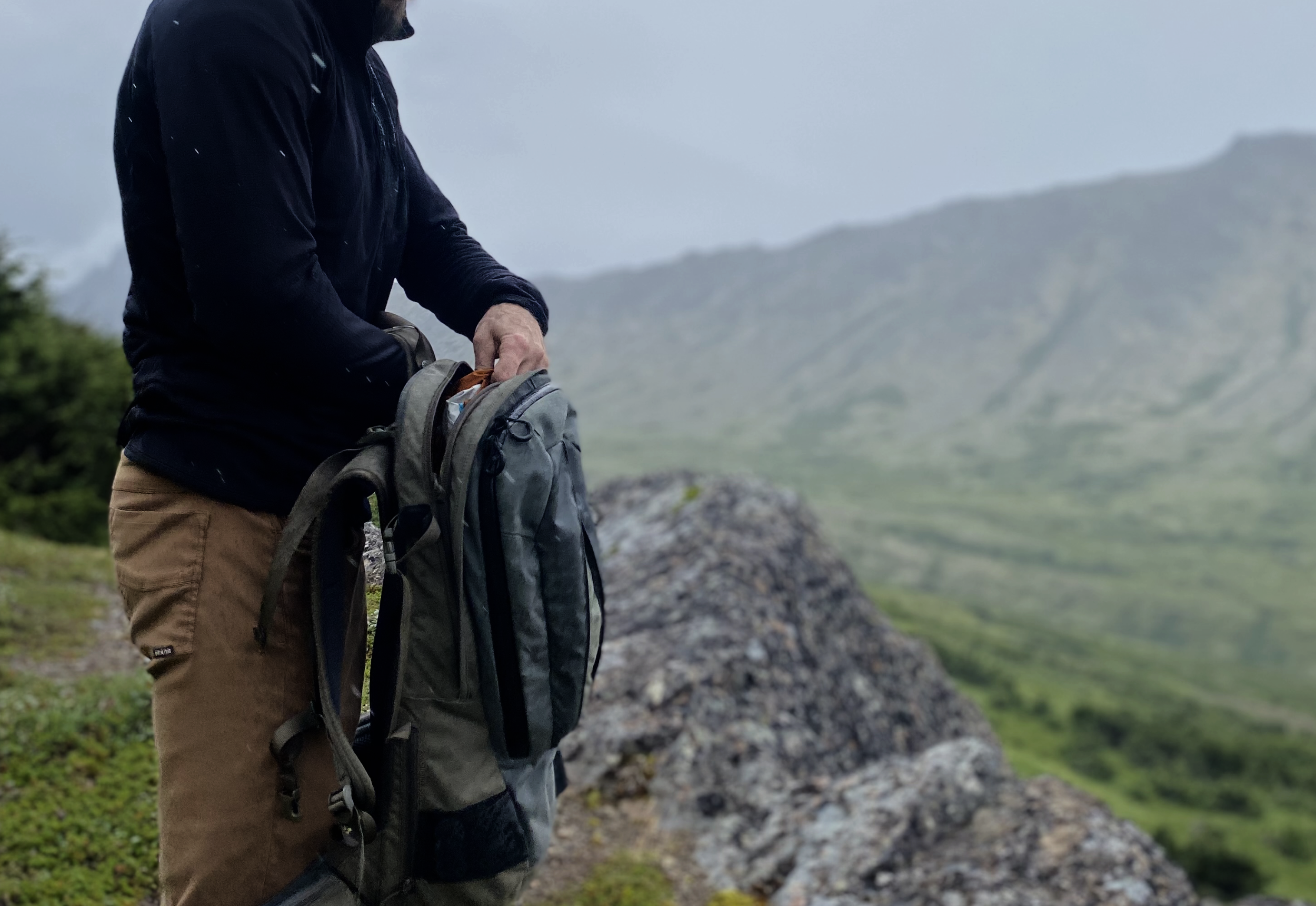 Person with a backpack standing on a rocky outcrop with a mountainous landscape in the background